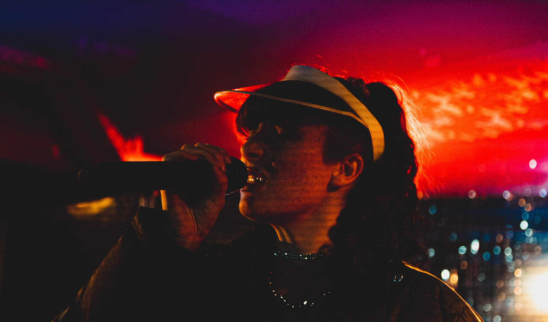 Person singing into a microphone wearing a visor cap and illuminated by warm red and orange stage lights.
