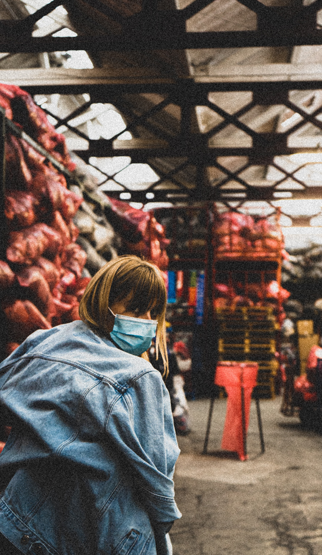 Person wearing a blue surgical mask and denim jacket in a warehouse filled with large bags stacked on shelves.