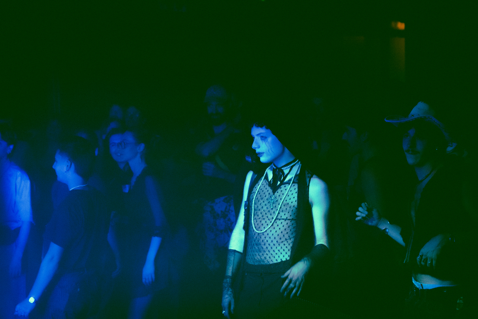 People dancing in a dimly lit club with blue and green lighting, featuring a person in a mesh top and pearl necklace.