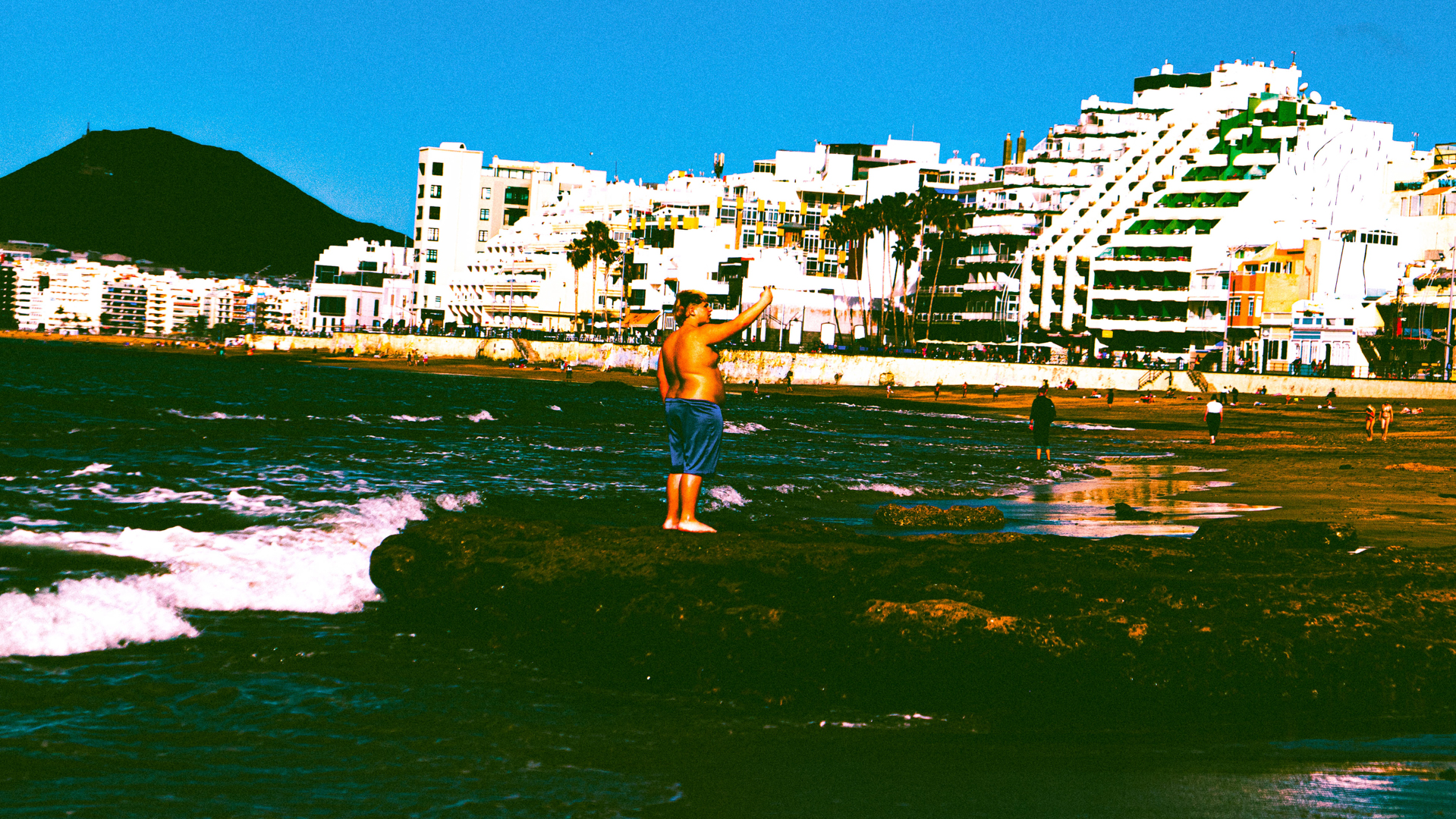 A boy in blue shorts stands on a rock near ocean waves with white buildings and a mountain in the background.