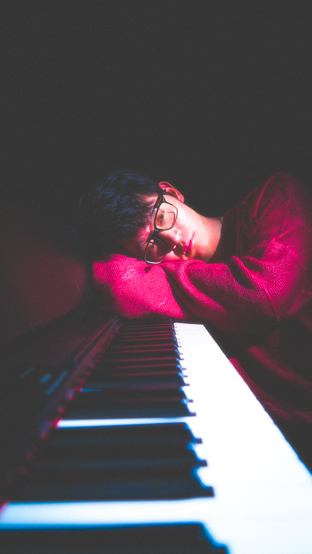 Young man in glasses resting his head on folded arms on a piano keyboard in low light.
