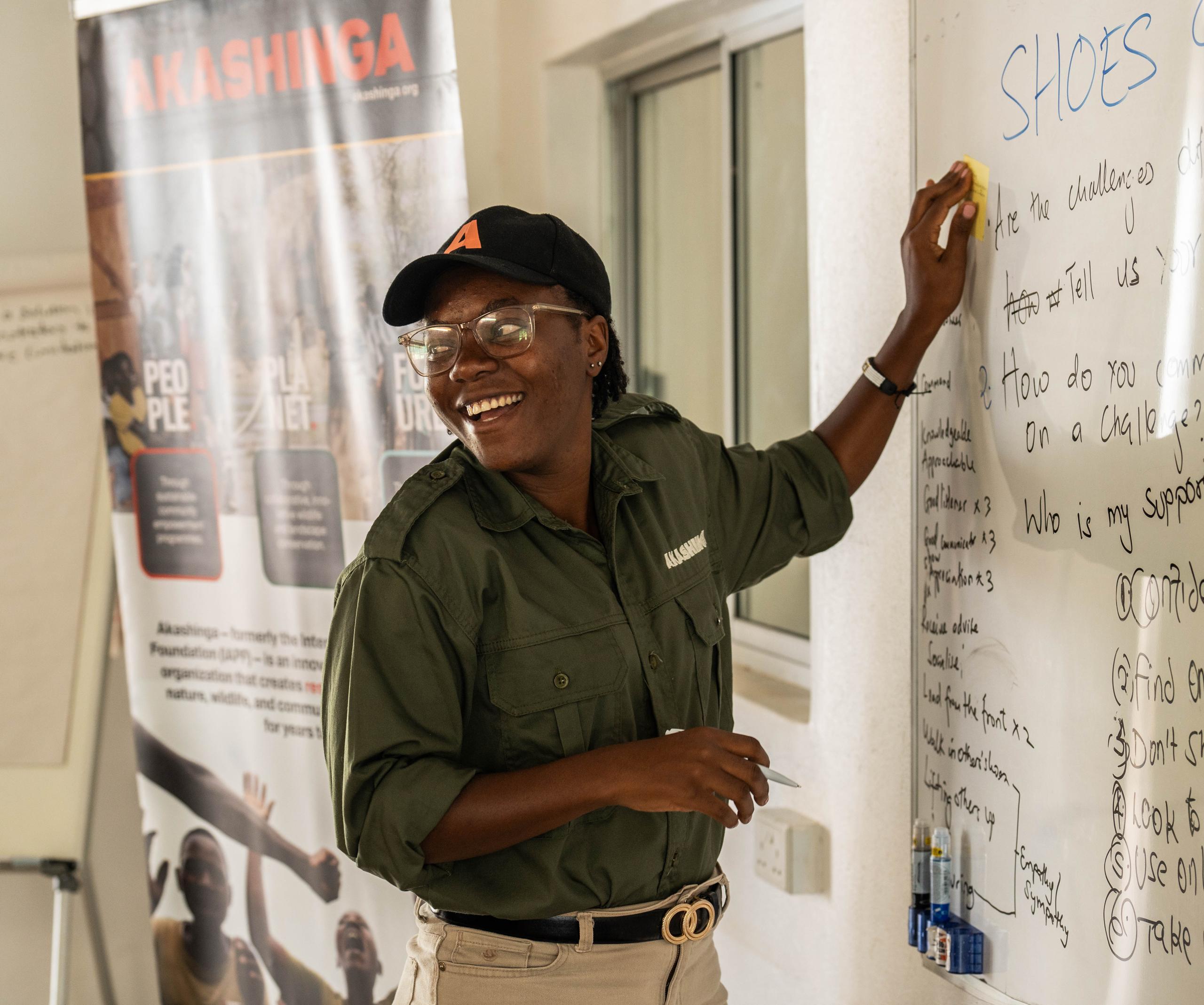 A woman wearing glasses, a hat, and a shirt with the Akashinga logo stands at a whiteboard, with her hand gesturing at the text written on it.