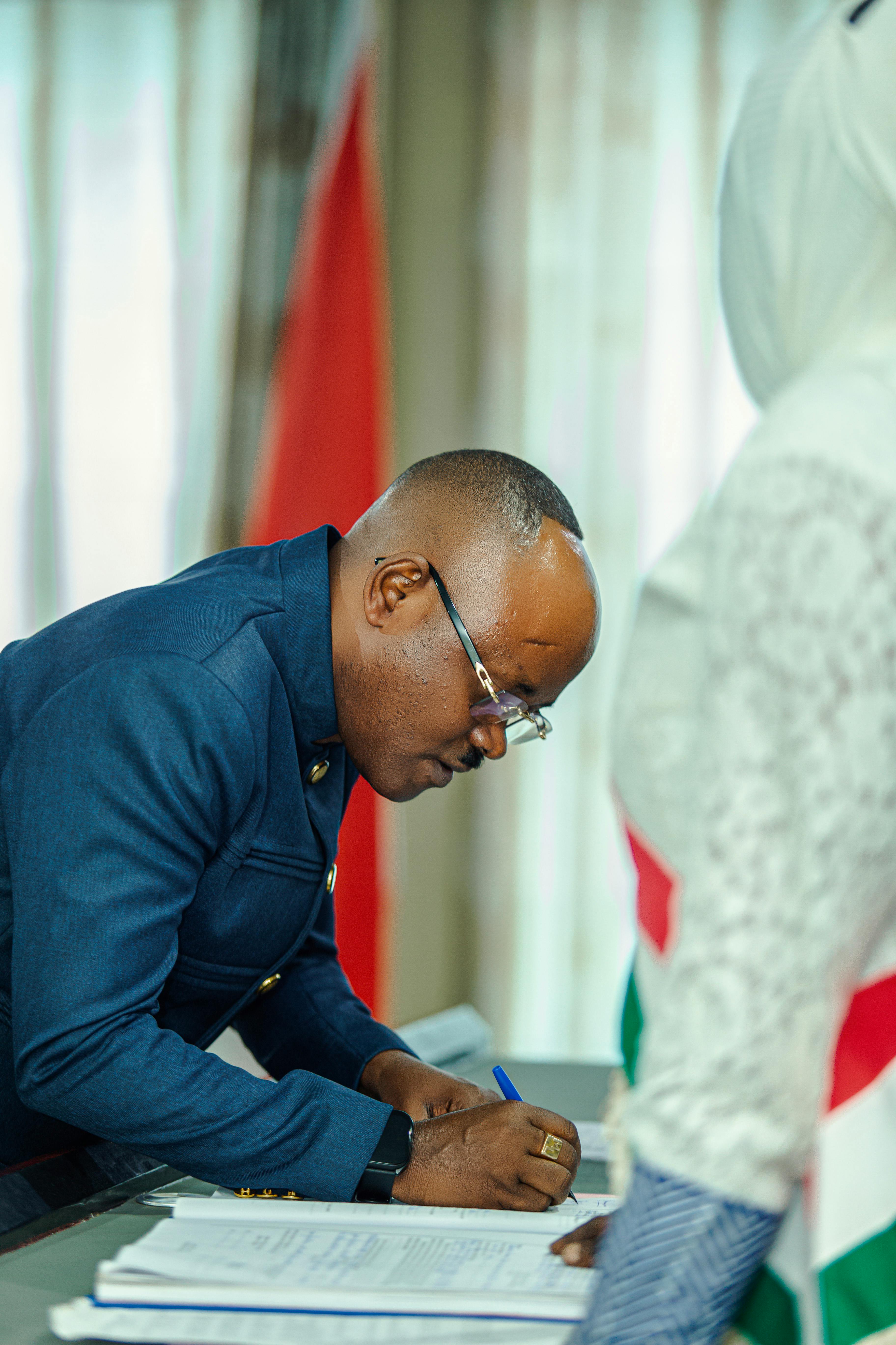 A man wearing glasses and a suit leans over a table and signs a document. In the background is an indistinct flag.
