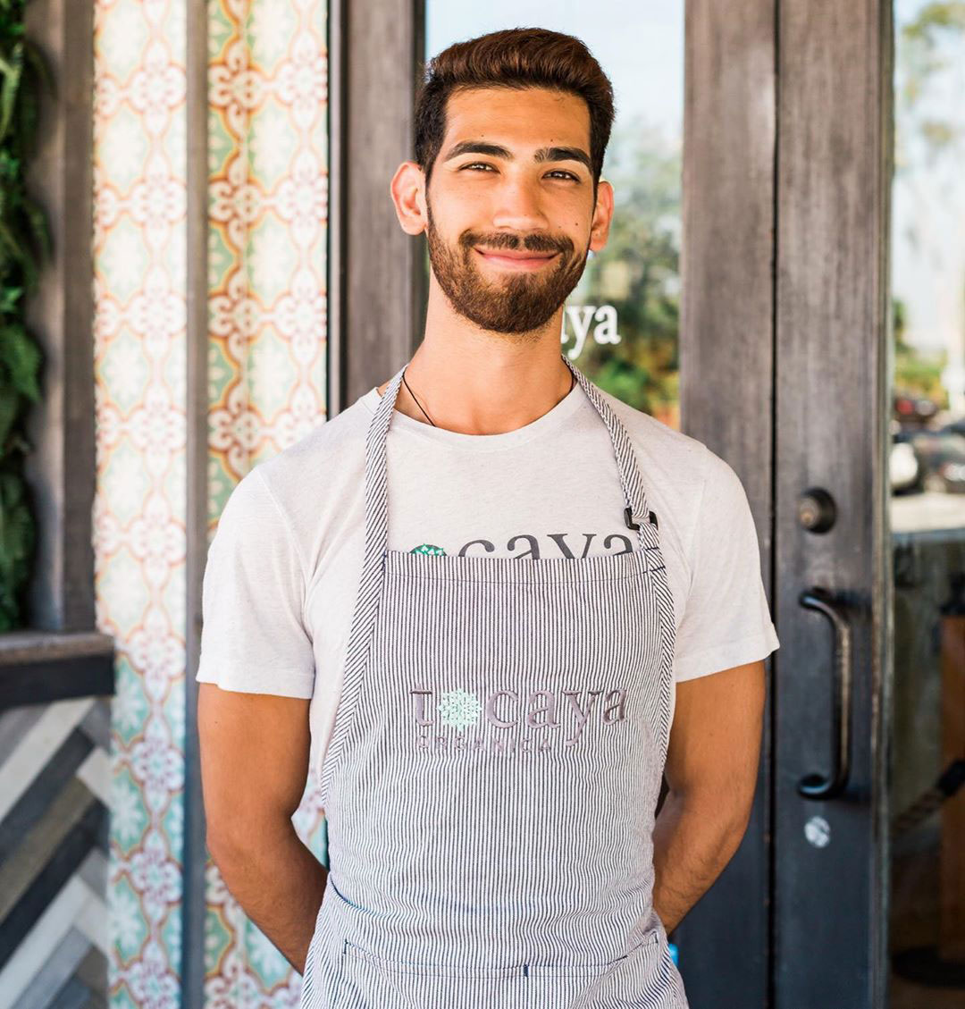 Smiling man with a beard wearing a white T-shirt and striped apron standing in front of a patterned wall and glass door.
