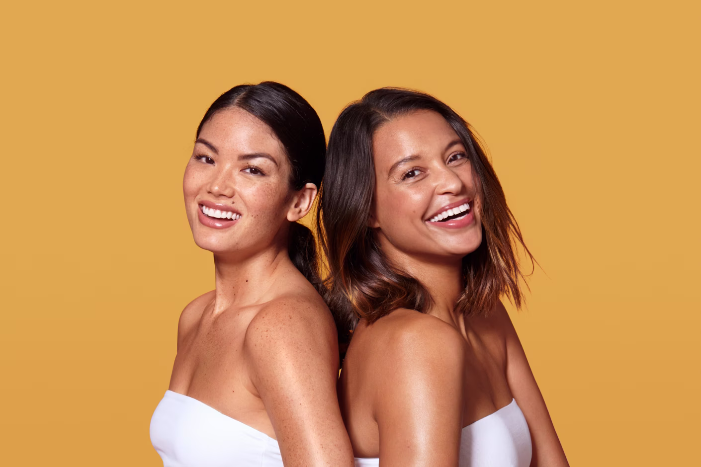 Two smiling women with bare shoulders standing back to back against a yellow background.