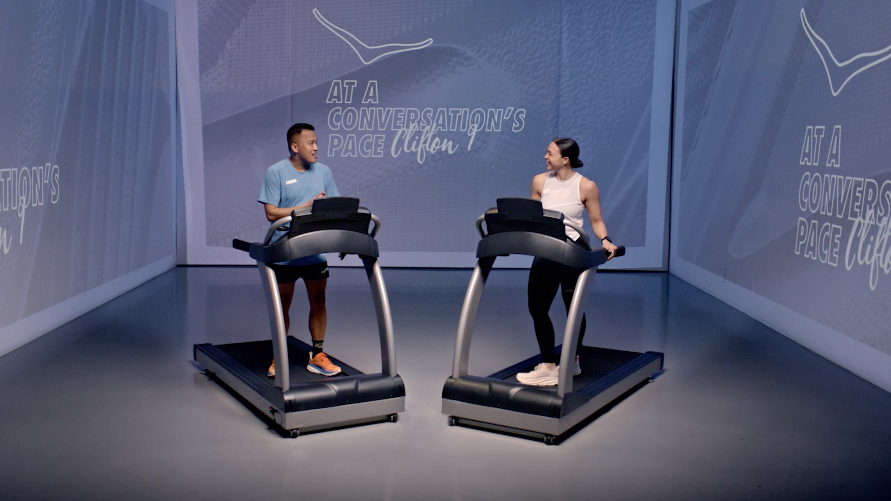 A man and woman smiling and talking while standing on adjacent treadmills in a minimalist room.