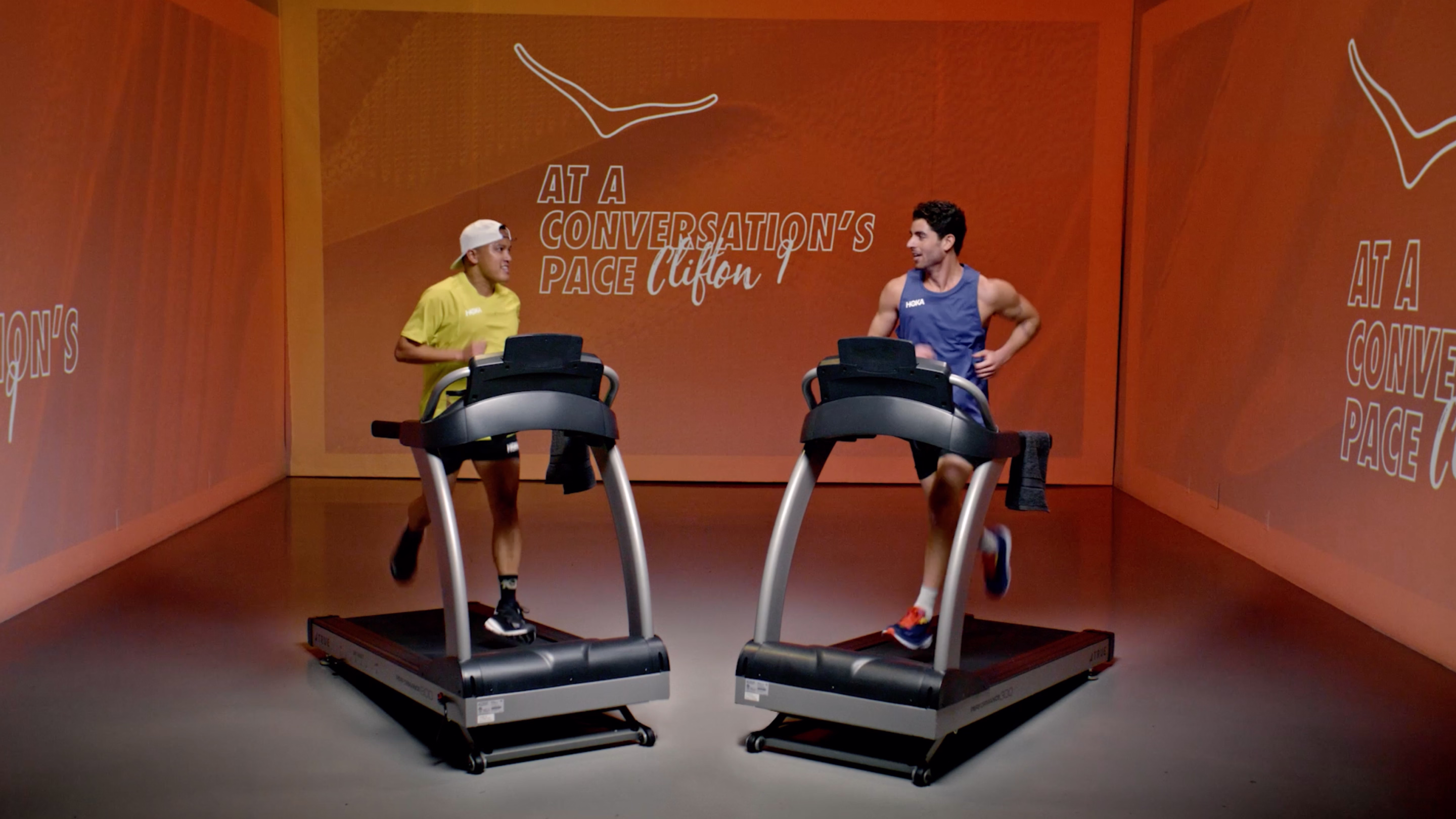 Two men running on treadmills facing each other with orange background displaying the text 'At a conversation's pace Clifton'.