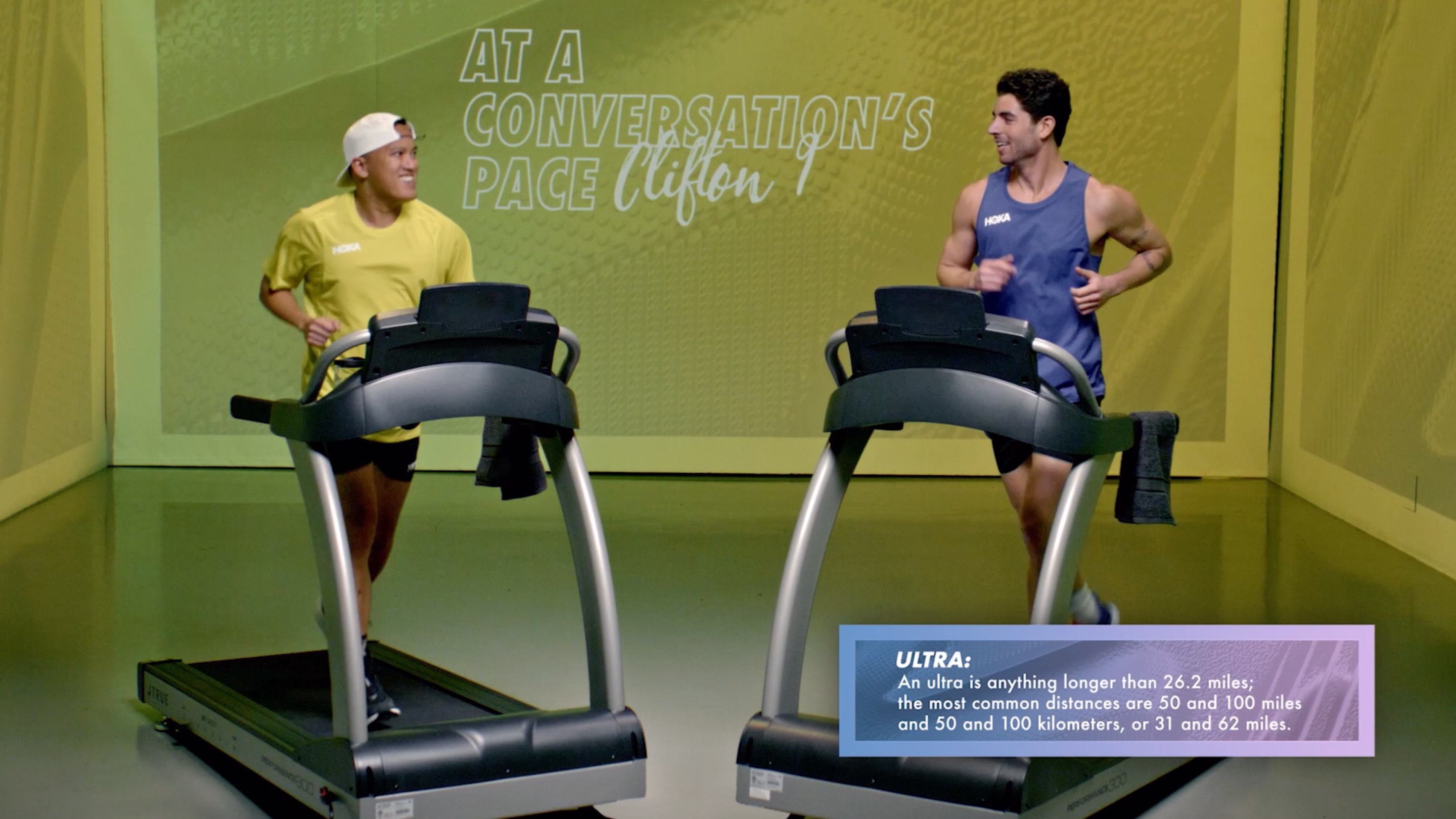 Two men running and smiling on treadmills in a studio with a green wall background displaying the text 'AT A CONVERSATION'S PACE' and a box explaining 'ULTRA: An ultra is anything longer than 26.2 miles; the most common distances are 50 and 100 miles and 50 and 100 kilometers, or 31 and 62 miles.'