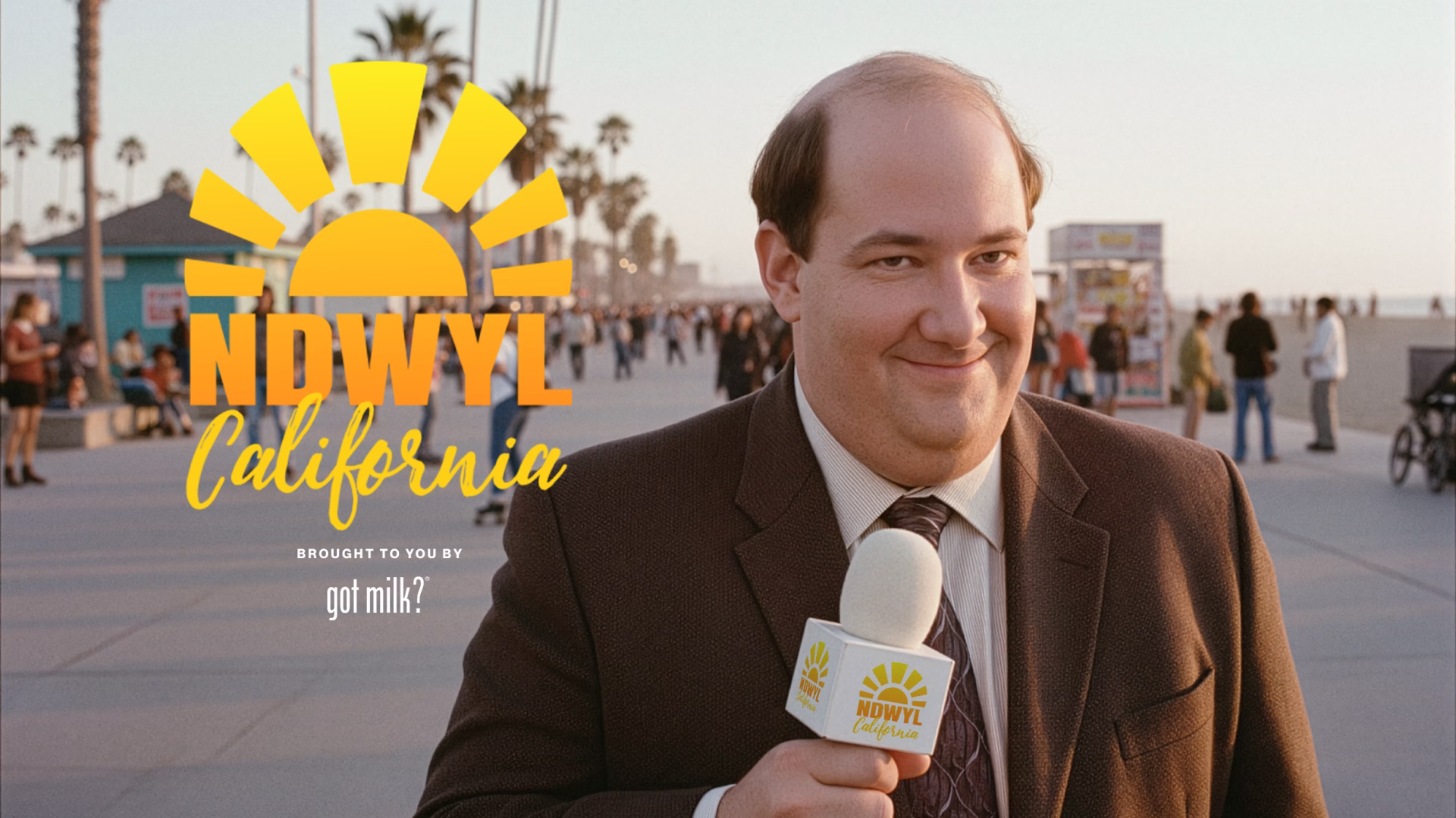 American actor Brian Baumgartner in a brown suit smiling and holding a microphone with the NDWYL California logo on a busy beachside boardwalk.