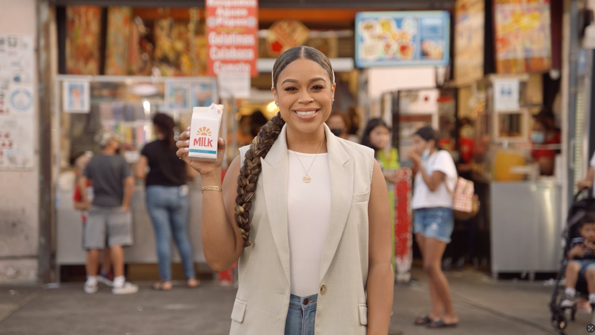 Actress Julissa Calderon smiling with long braided hair holding a carton of milk in front of a busy storefront.