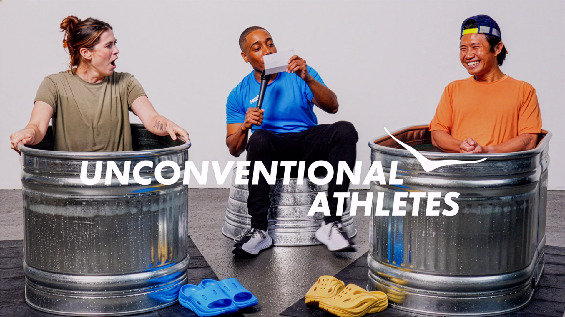 Two people sitting in metal tubs filled with ice water on either side of a man in a blue shirt holding a microphone and paper, with the text 'Unconventional Athletes' overlaid.