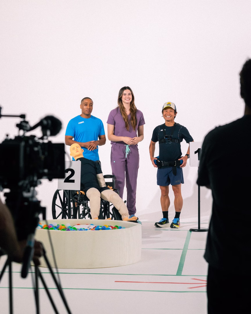 Three people standing in a studio setup with one person in scrubs, another in athletic gear, and a mannequin sitting in a wheelchair in front of numbered signs 1 and 2.