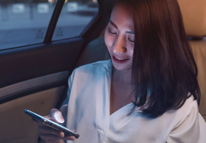 A woman, sitting in the back of a car, looks down at her mobile phone smiling.