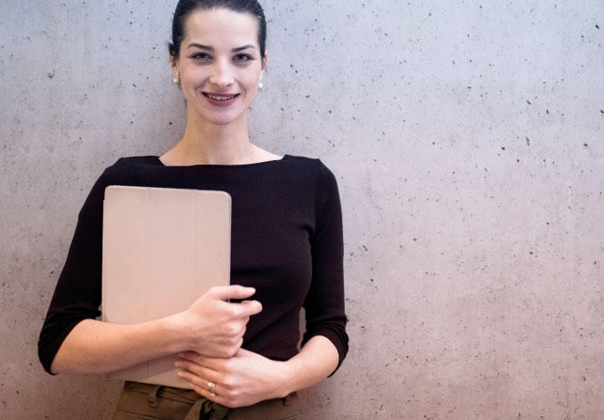 Woman smailing, standing against wall holding laptop.
