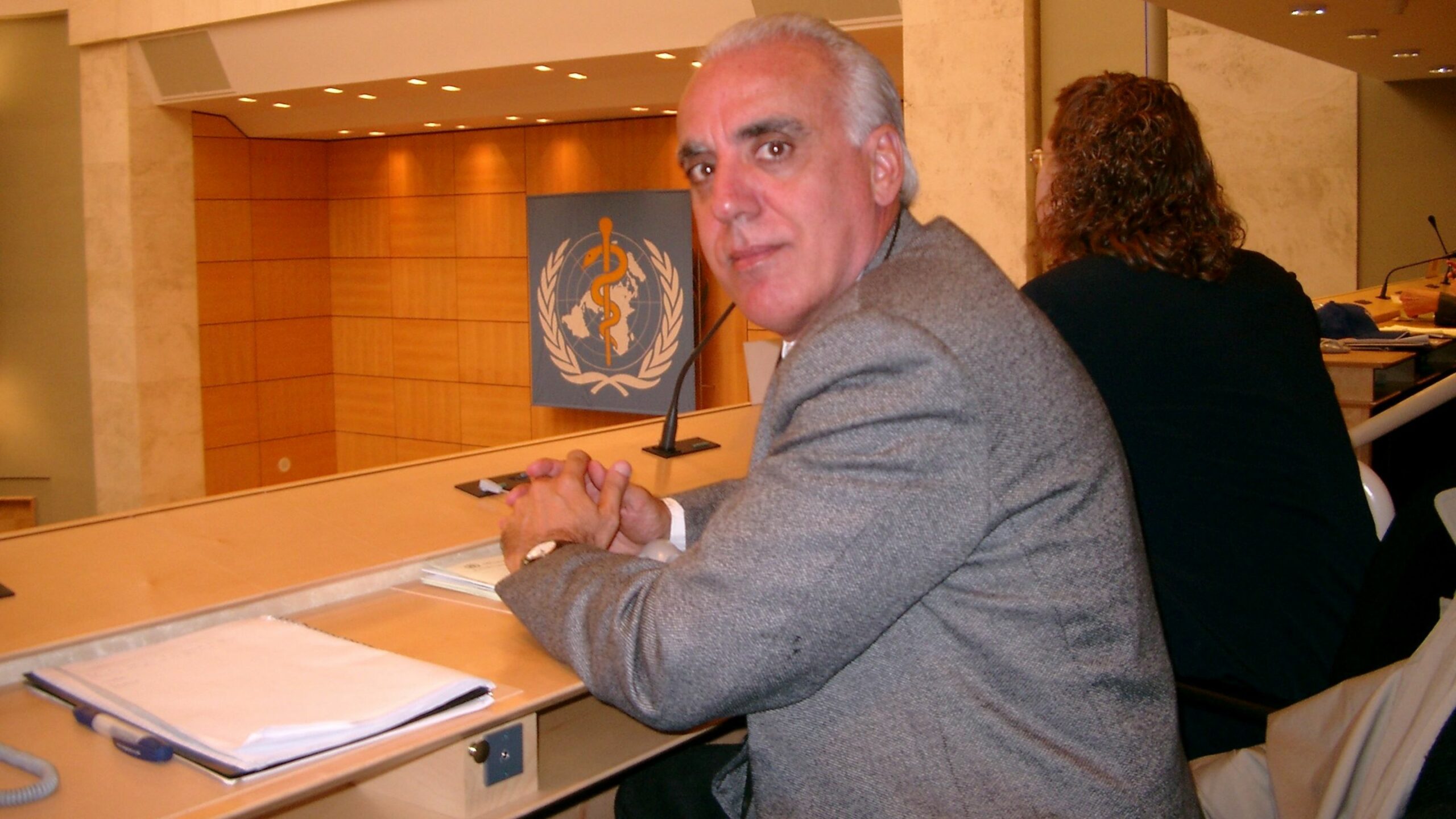 A person is seated in a conference room with a WHO emblem in the background.