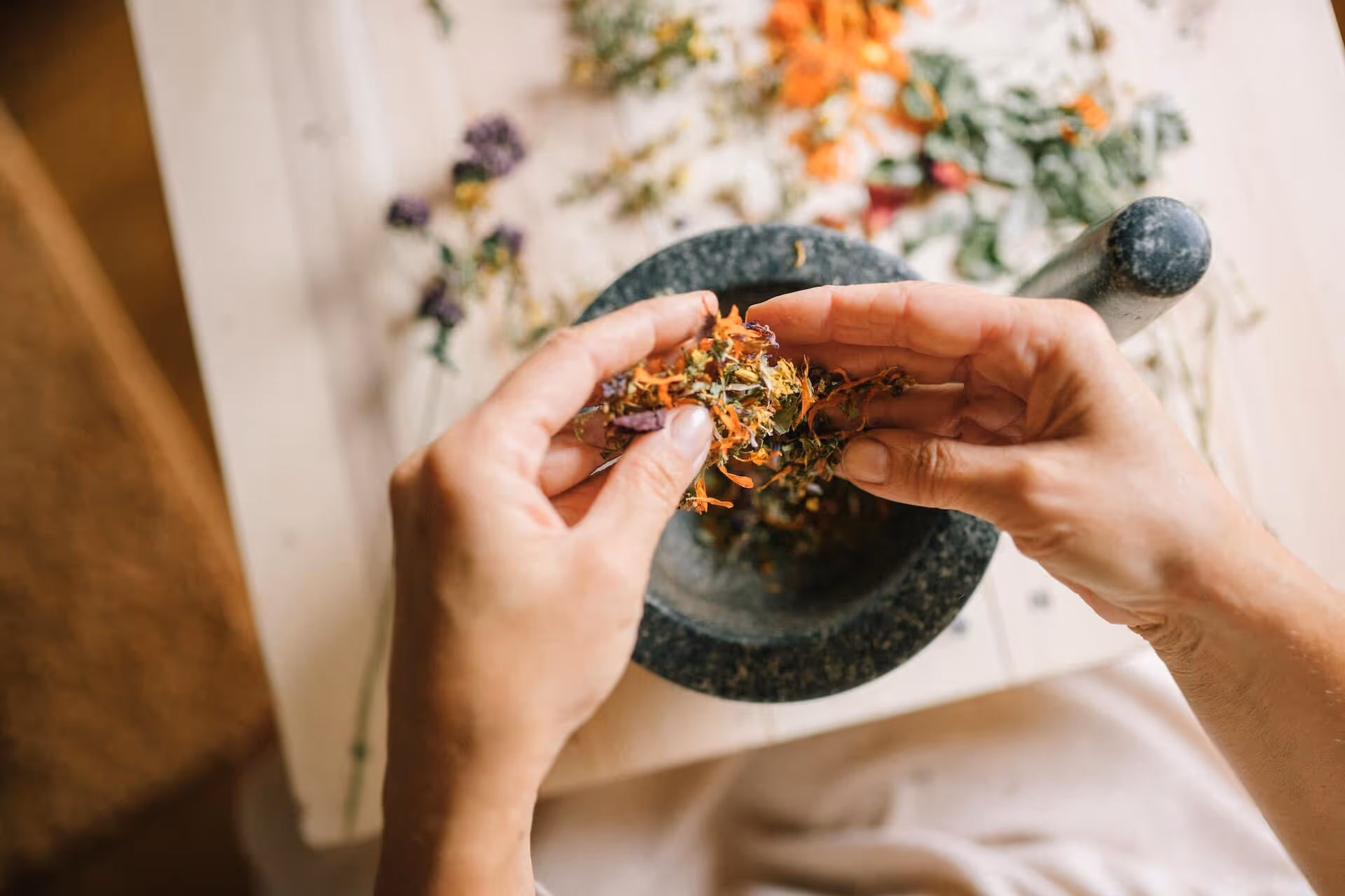 Hands placing dried flowers into a stone mortar with a pestle on a wooden surface.