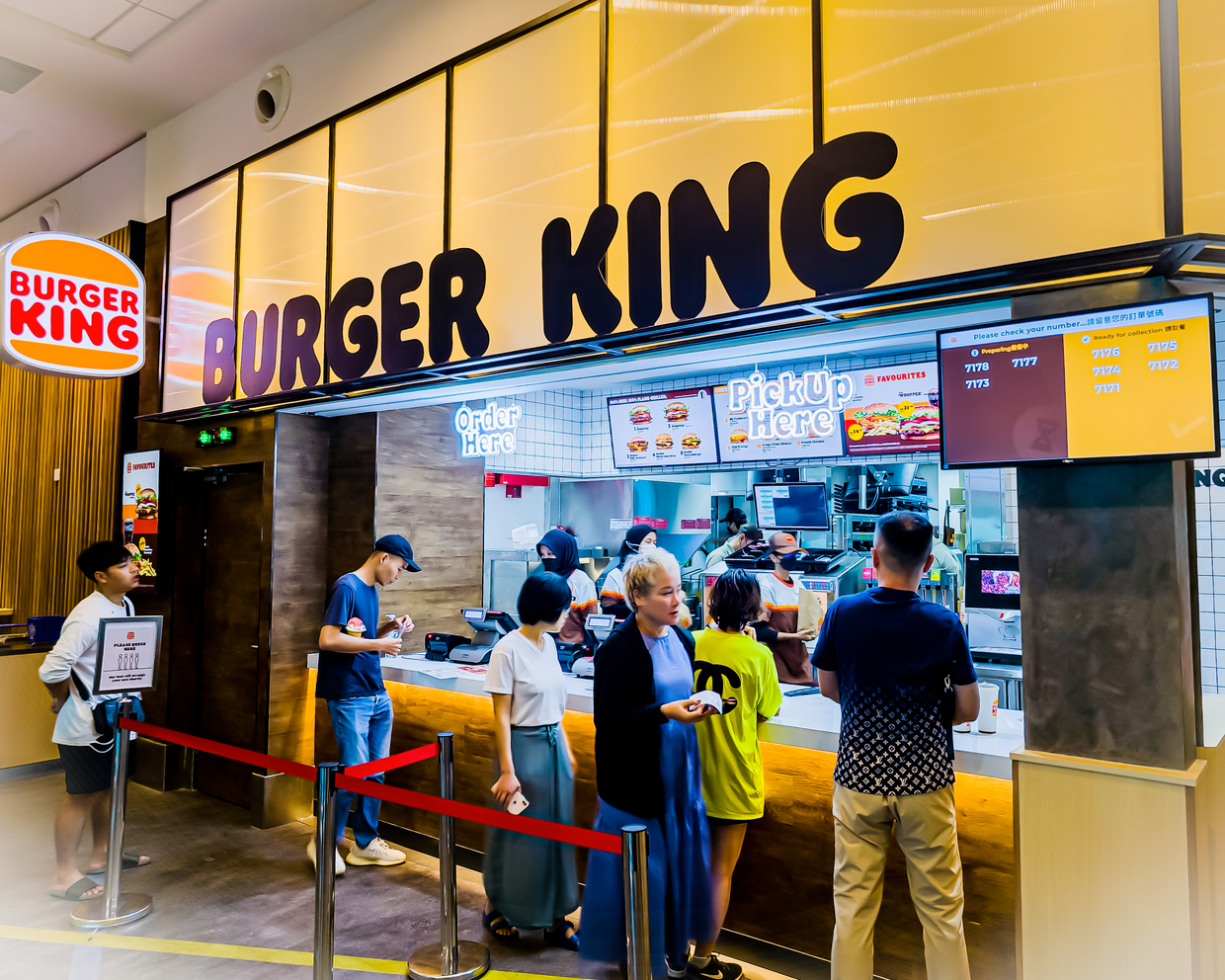 Customers waiting in line and ordering at a Burger King fast-food counter with illuminated 'Order Here' and 'Pick Up Here' signs.