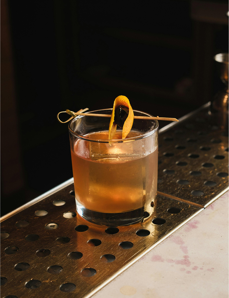 Old fashioned cocktail with an orange peel and cherry garnish on a perforated metal bar surface.