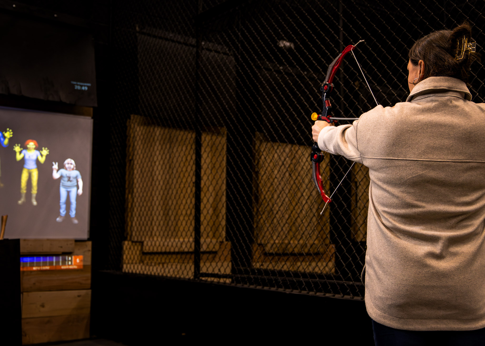 Person aiming a small red bow and arrow at an indoor target area behind a chain-link fence with animated characters displayed on a nearby screen.