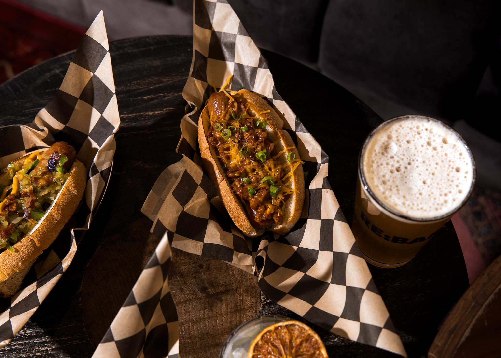 Two hot dogs with toppings in checkered paper baskets on a dark wooden table, next to a pint of beer with foam.