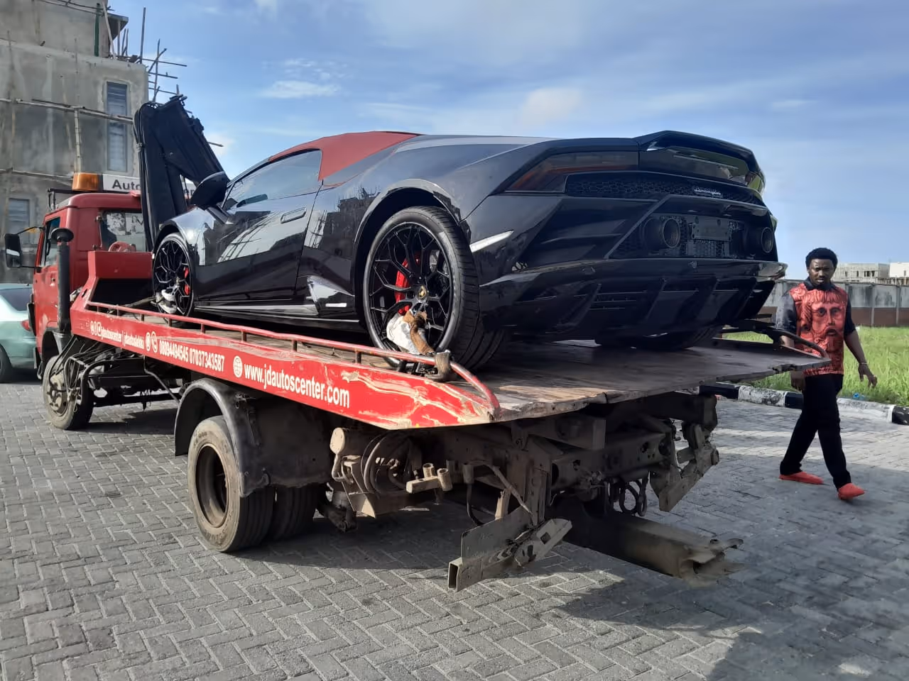 Black sports car with red roof on the back of a red flatbed tow truck on a paved area with a man walking nearby.