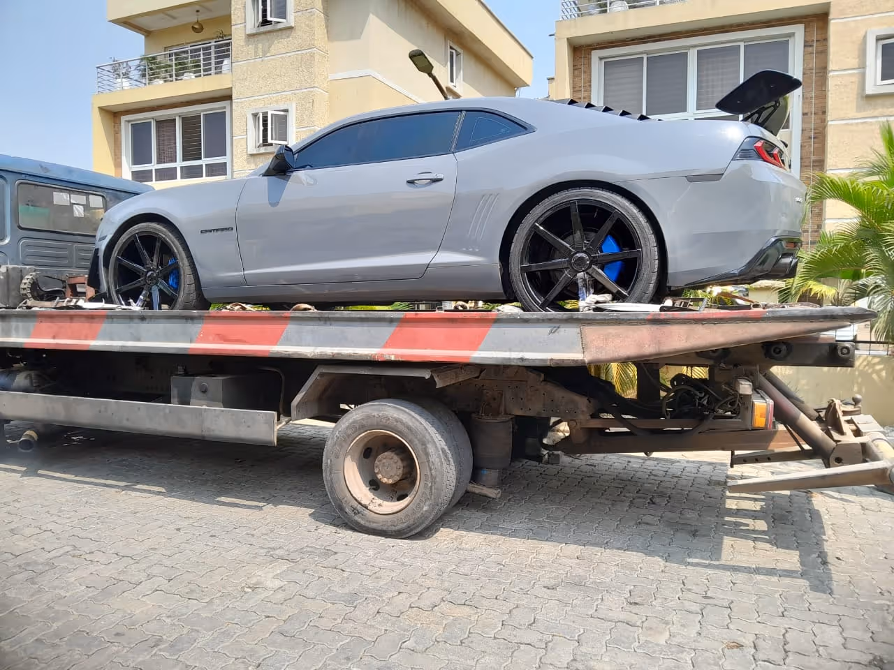 Silver sports car with black rims and blue brake calipers on a flatbed tow truck in front of beige residential buildings.