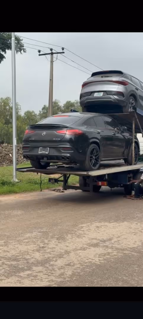 Two cars, one black Mercedes-Benz and one silver vehicle, loaded on a car carrier trailer parked on a roadside.