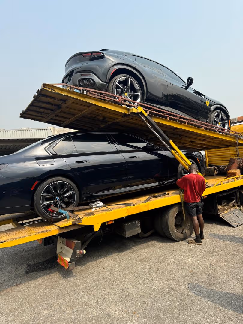 Two black luxury cars, a BMW sedan below and a Ferrari SUV above, loaded on a yellow tow truck with a man standing nearby.