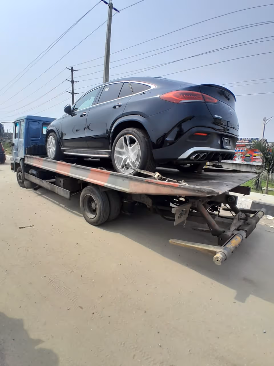 Black Mercedes-Benz SUV loaded on the back of a flatbed tow truck on a dirt road.