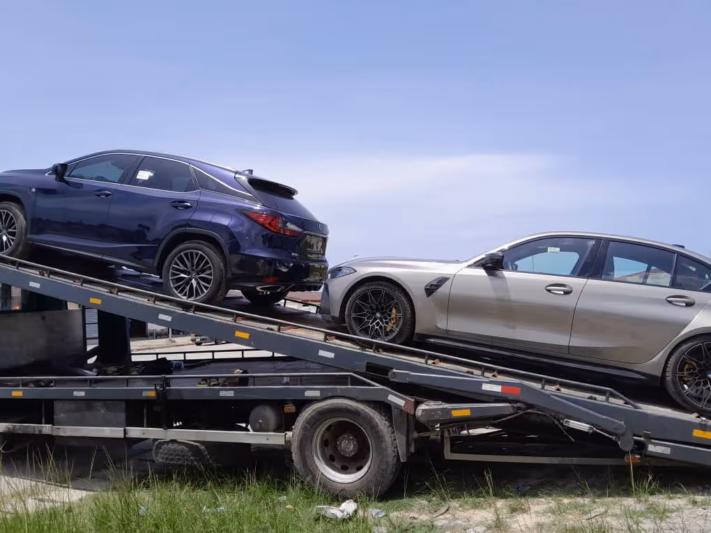 Two luxury cars, a dark blue SUV and a silver sedan, being loaded onto a car carrier truck on a grassy roadside.