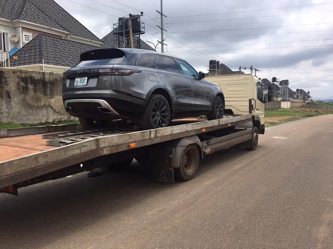 Gray Range Rover SUV loaded on a flatbed tow truck parked on the side of a paved road with houses and cloudy sky in the background.