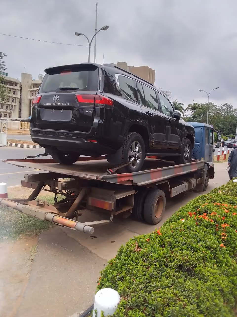 Black Toyota Land Cruiser VX loaded on a flatbed tow truck near a sidewalk with bushes and buildings in the background.