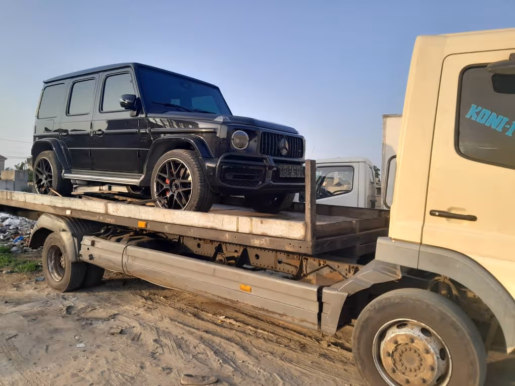 Black Mercedes-Benz G-Class SUV loaded on the back of a flatbed tow truck on a dirt surface.