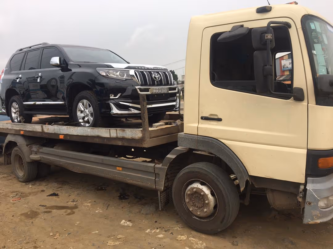 Black Toyota Land Cruiser Prado SUV loaded on a flatbed tow truck on a dirt surface.