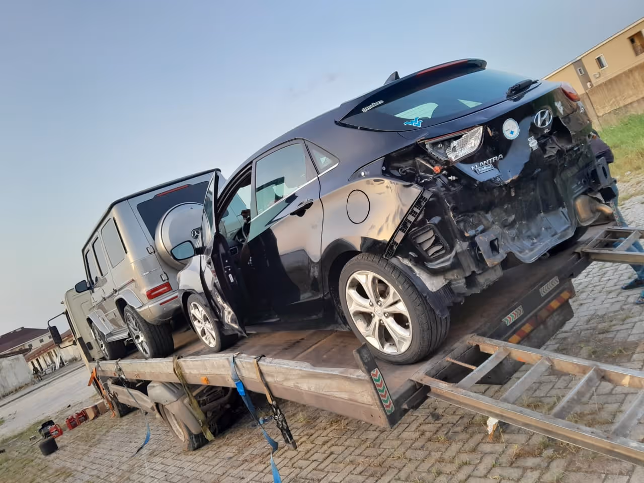 Damaged black Hyundai Elantra and gray SUV loaded on a tow truck ramp on a paved surface in an outdoor setting.