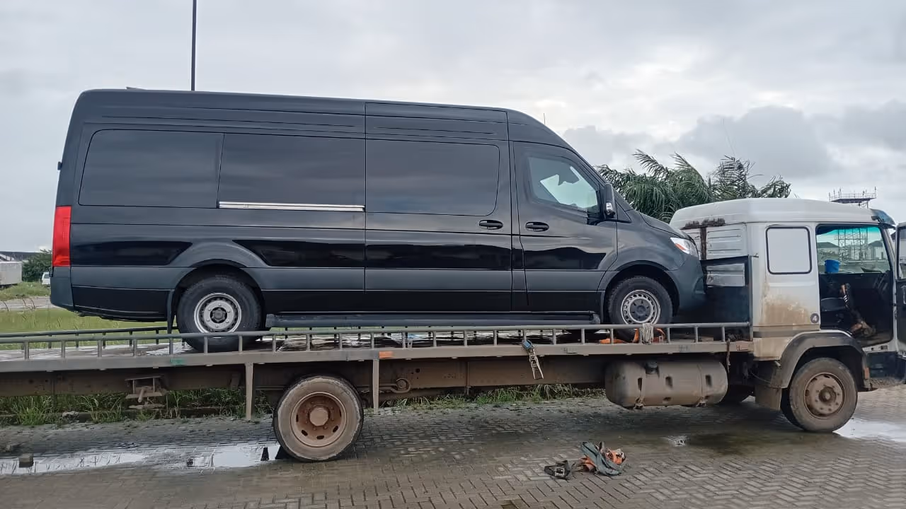 Black cargo van loaded on the back of a flatbed tow truck parked on a paved area on a cloudy day.