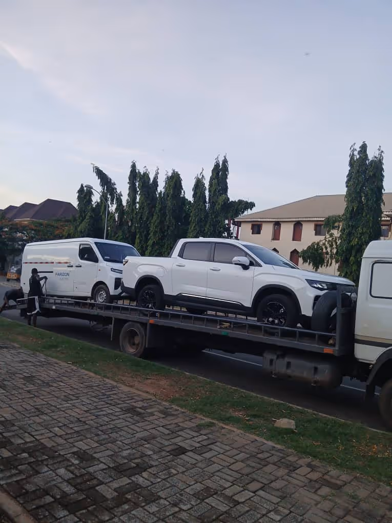 White pickup truck and white van loaded on a flatbed tow truck on a street with trees and buildings in the background.