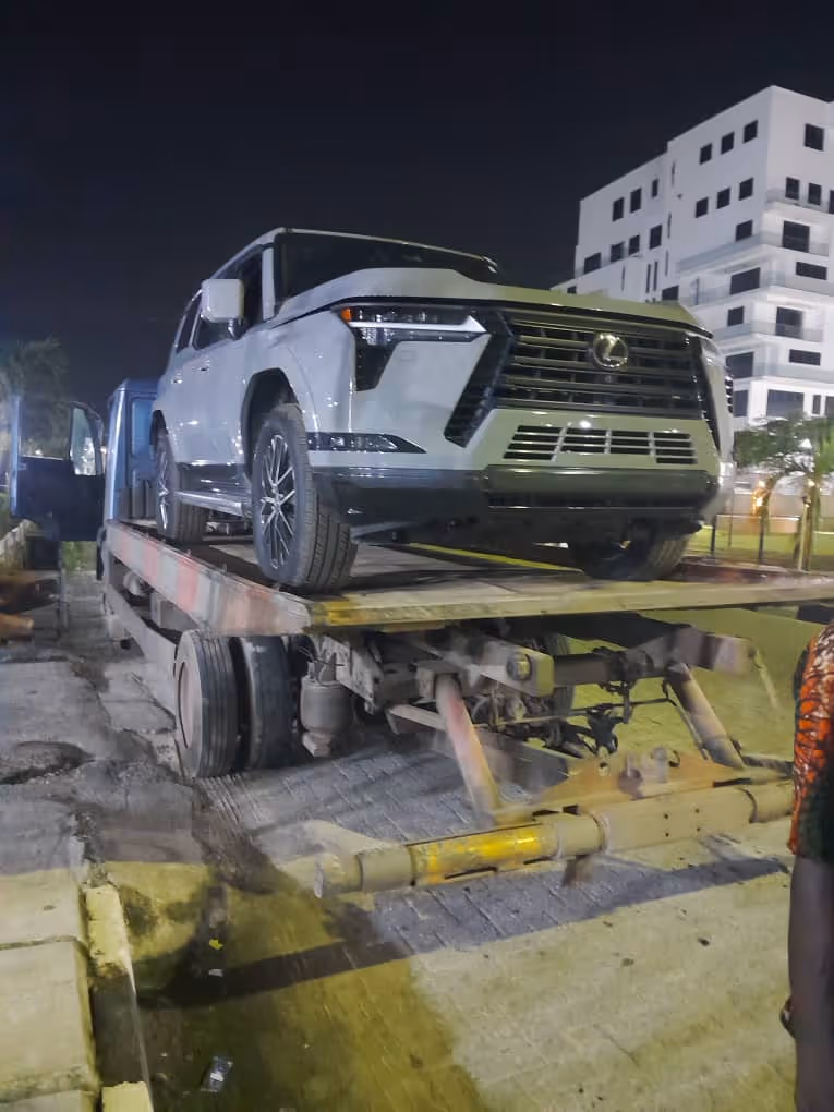 White Lexus SUV loaded on a flatbed tow truck at night on a paved street near modern buildings.