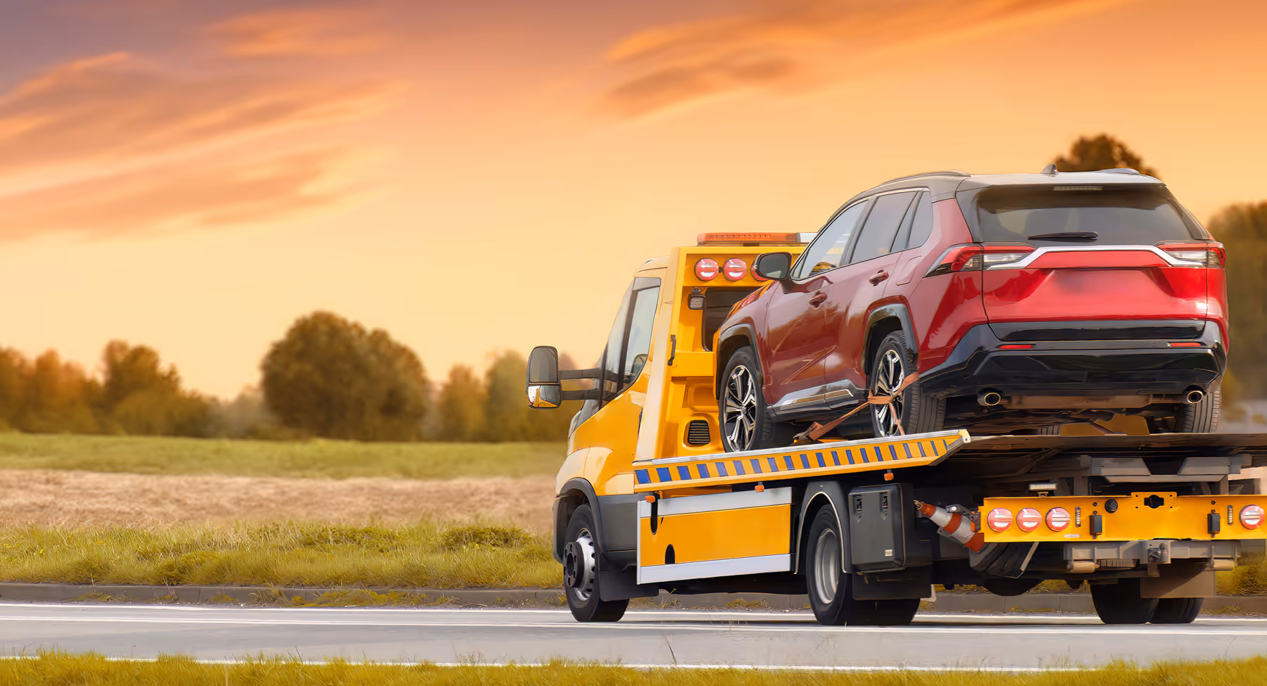 Yellow tow truck carrying a red SUV on its flatbed on a rural road at sunset.