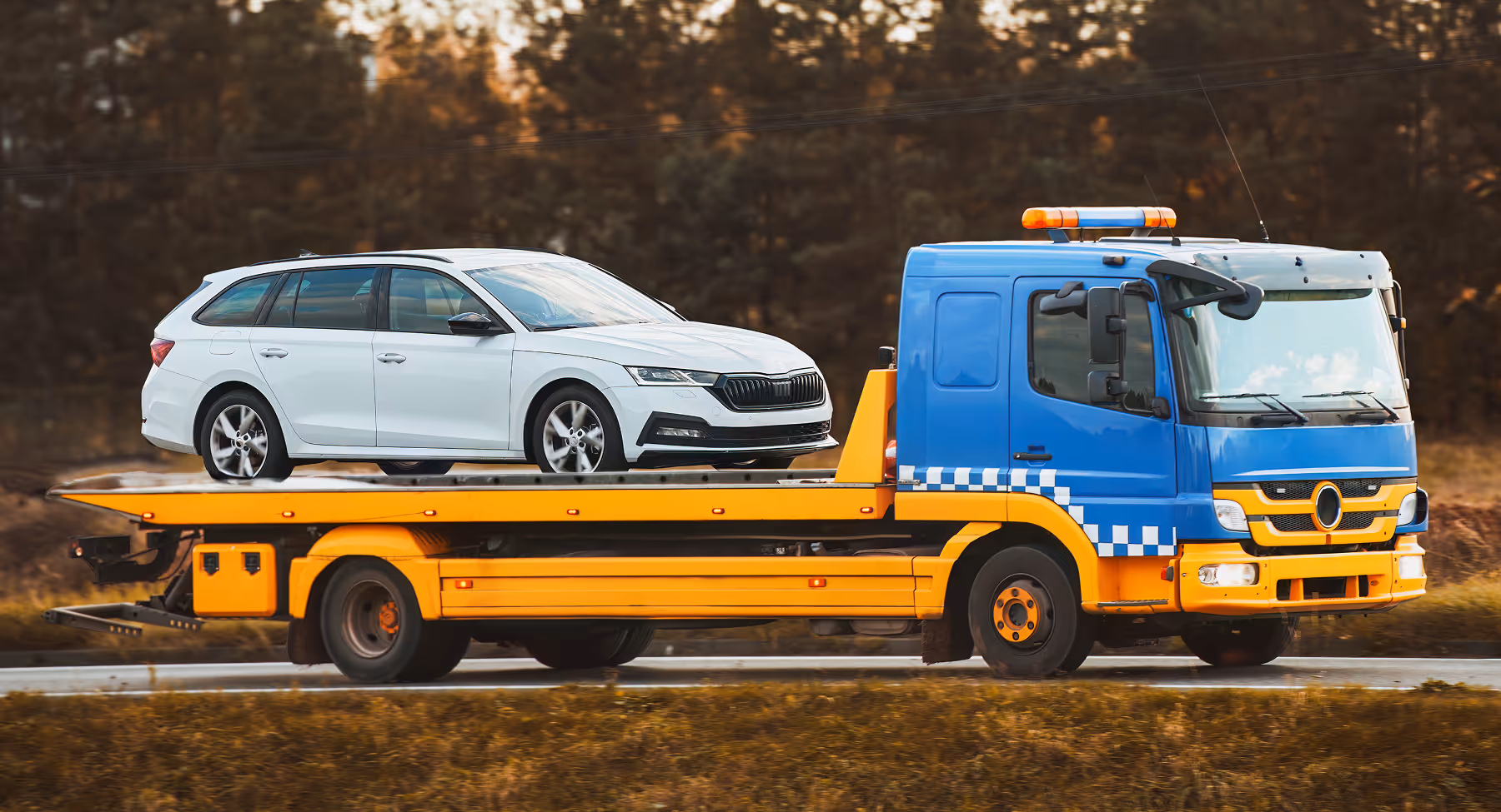 Blue and yellow tow truck carrying a white sedan car on a flatbed on a road.