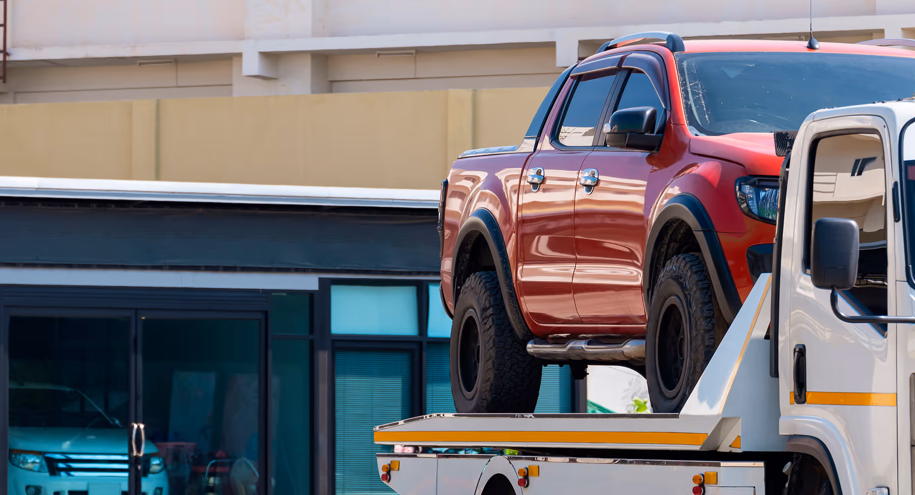 Red pickup truck loaded on the back of a white flatbed tow truck in front of a building with large windows.