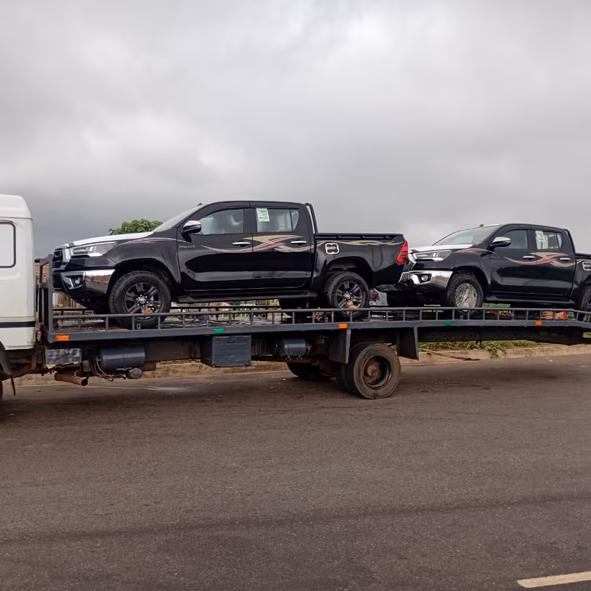 Two black pickup trucks with decals loaded on a flatbed tow truck on a road under cloudy sky.