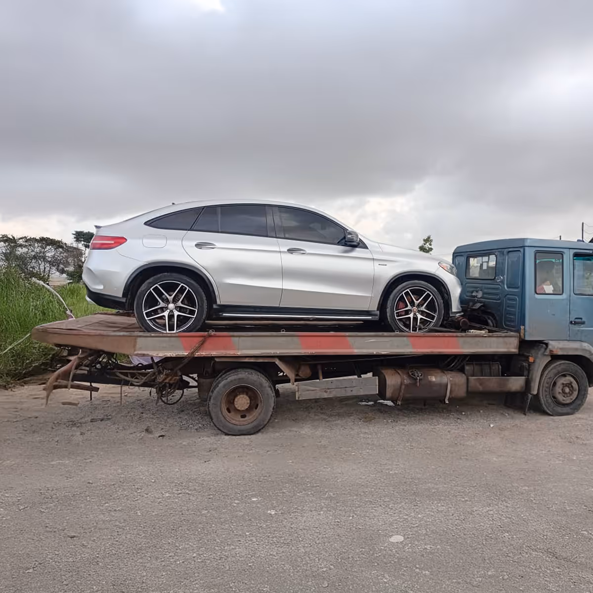 Silver SUV loaded on a flatbed tow truck under cloudy sky on a gravel road.