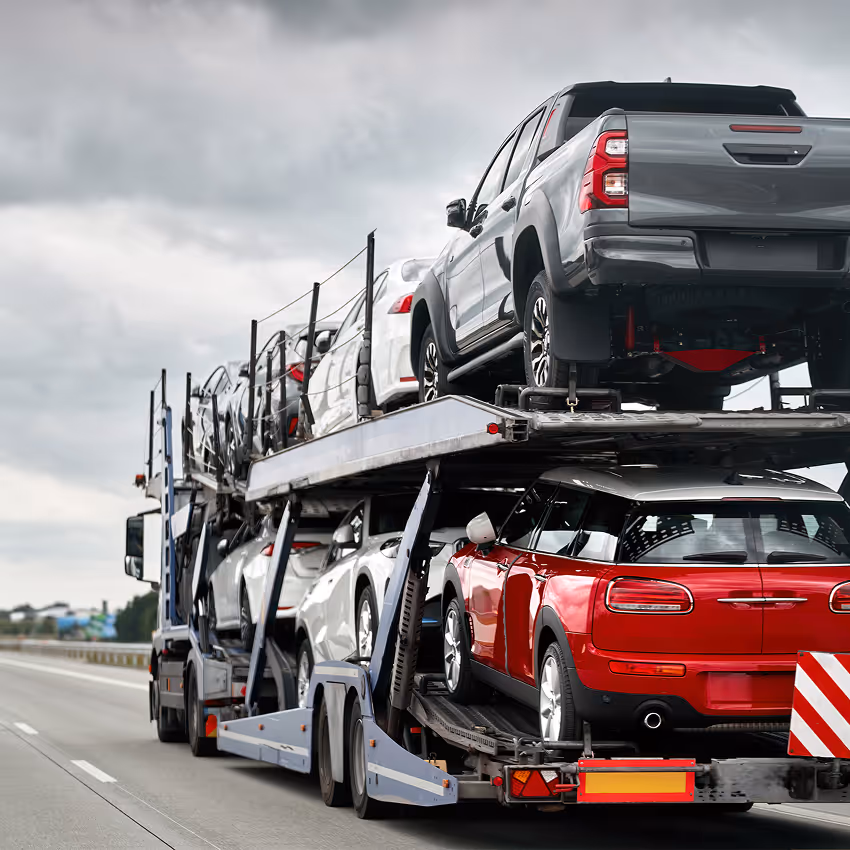 Car transporter trailer carrying multiple new cars on a highway under a cloudy sky.