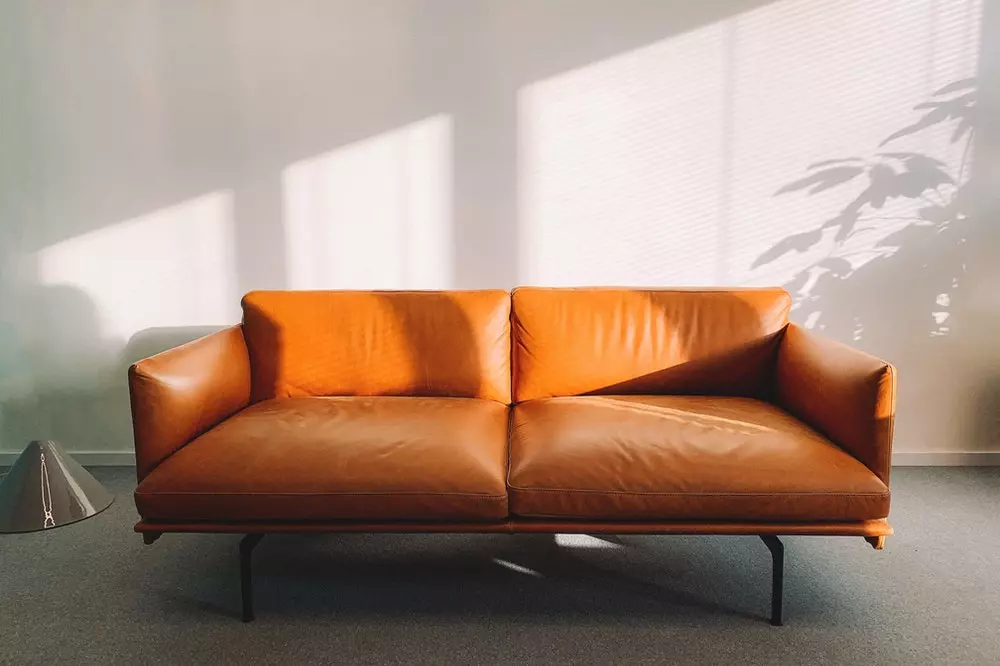Modern orange leather sofa illuminated by sunlight casting window shadows on a plain wall.