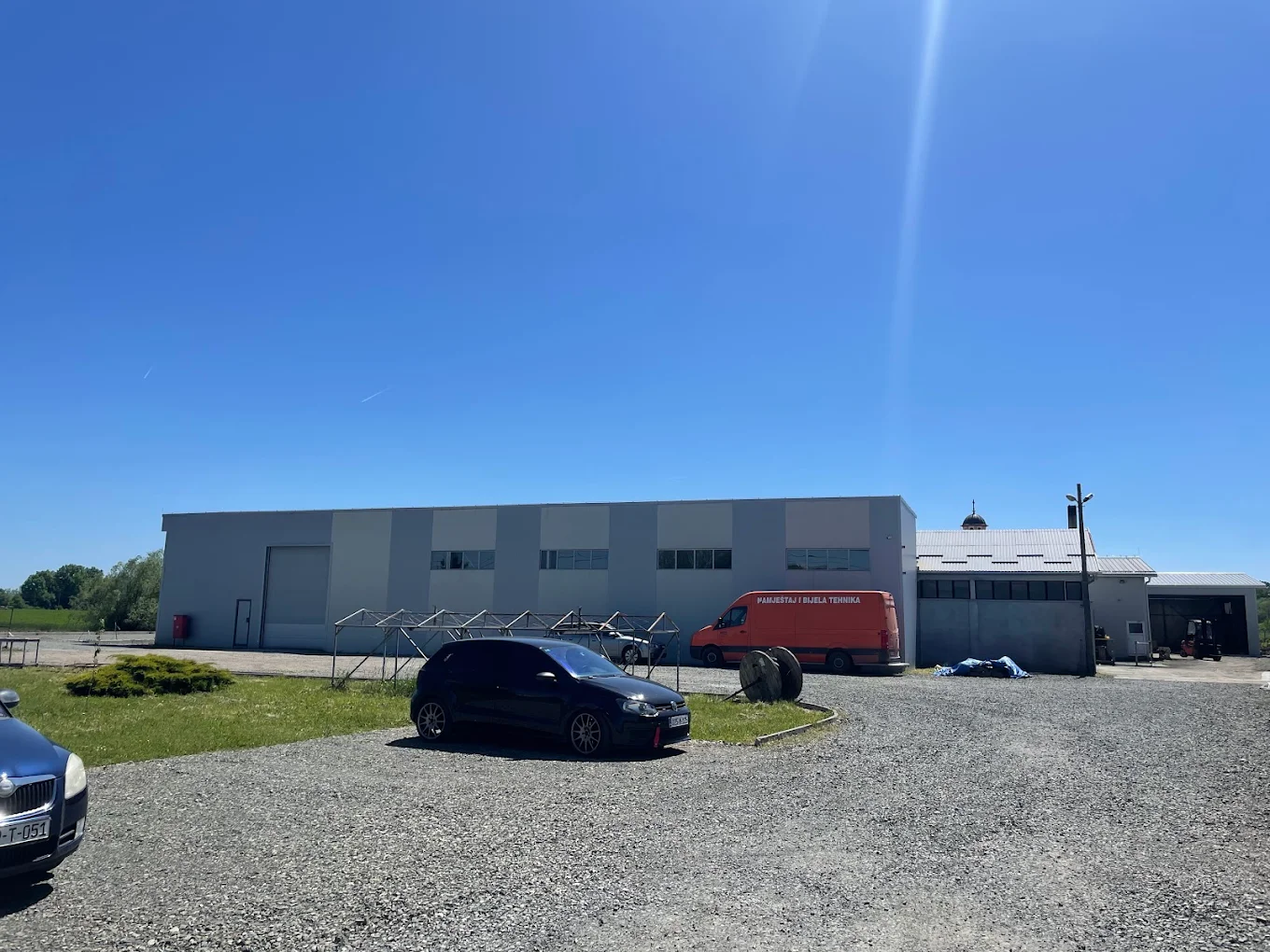 Industrial building with several parked vehicles including a black car and an orange van under clear blue sky.