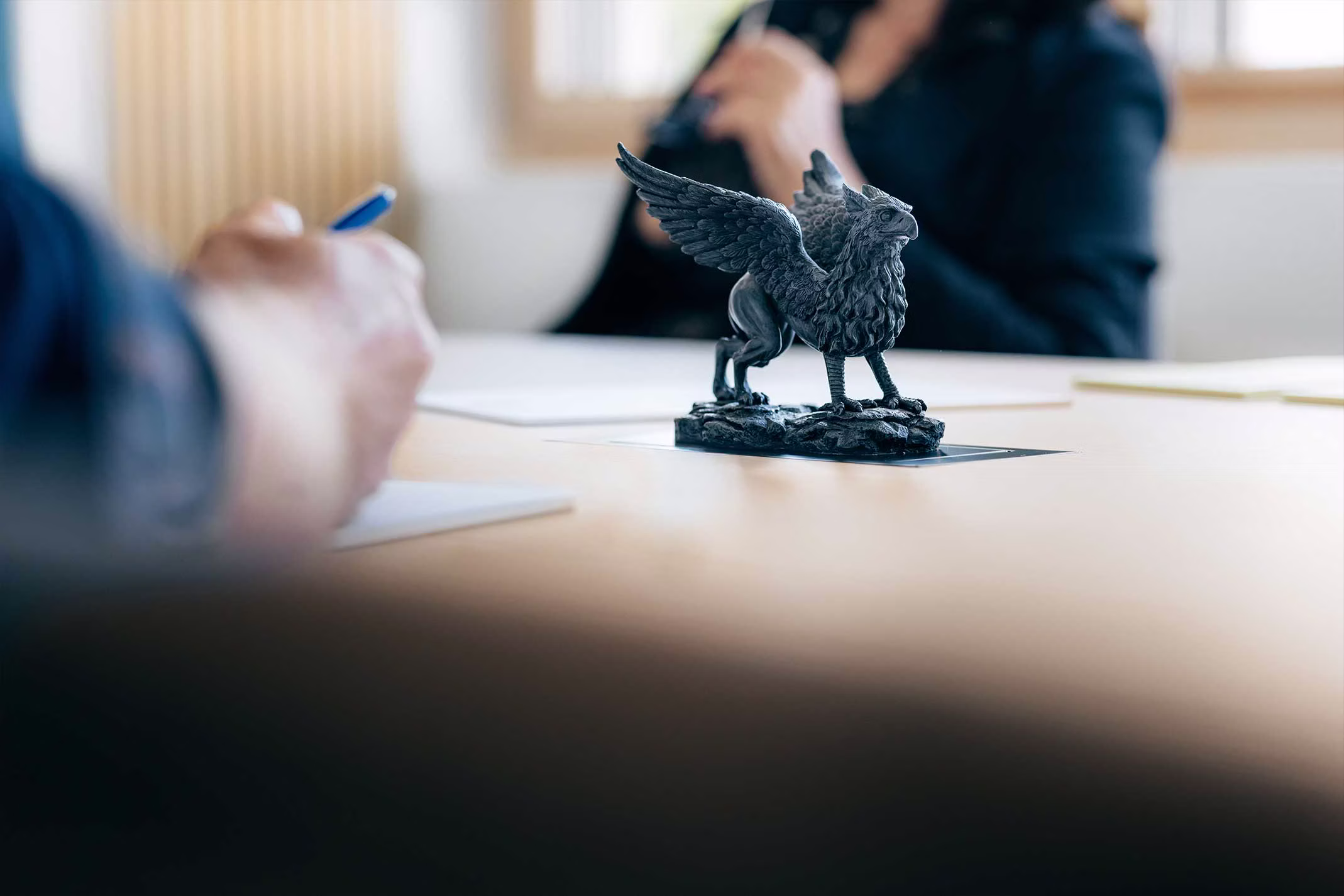 Griffin Trust – Vermögensschutz: Greif-Skulptur auf Konferenztisch, Beratung im Hintergrund – Symbol für Sicherheit und werterhaltende Strategien.