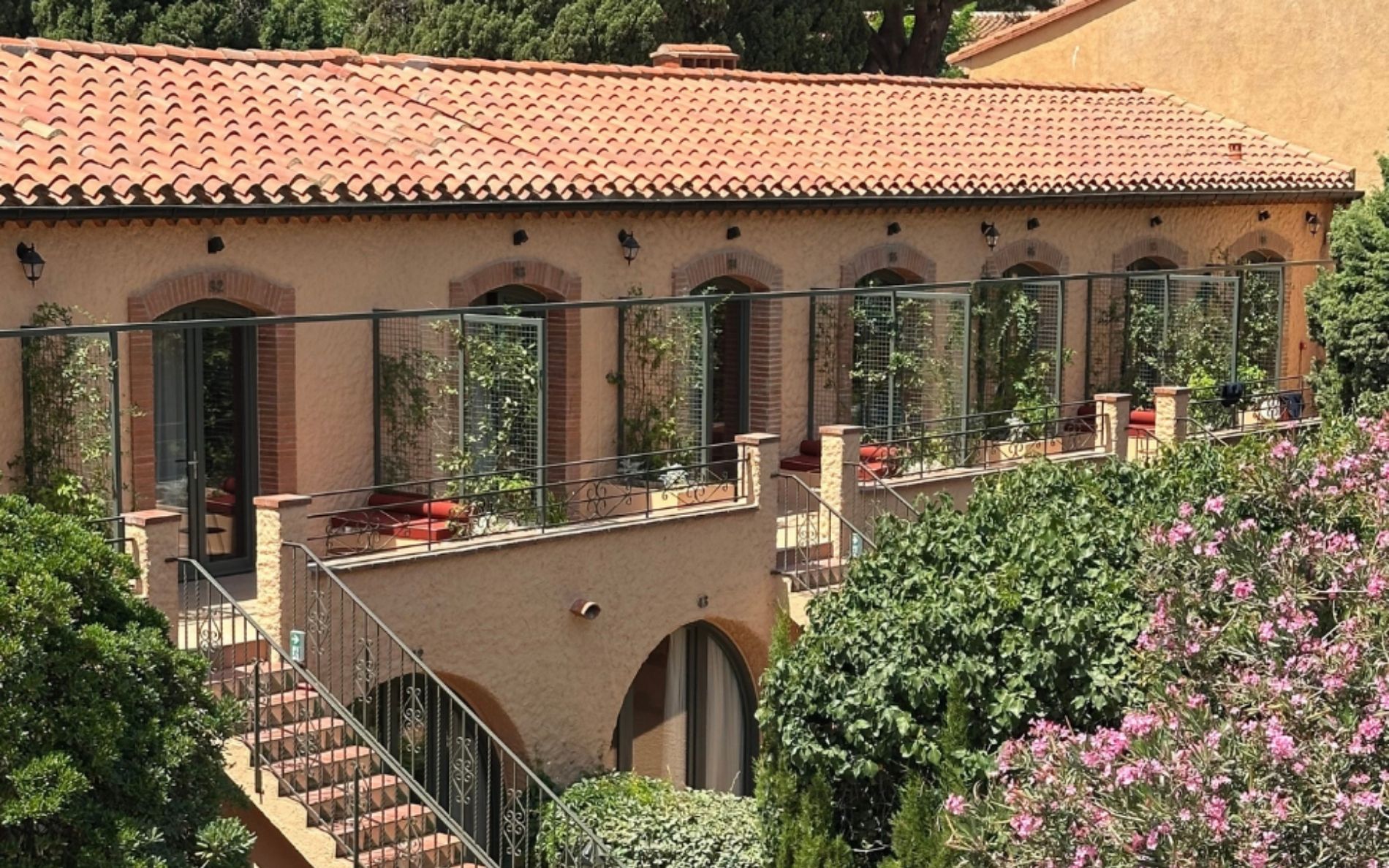 Mediterranean-style building with tiled roof, arched doors, balcony plants, and surrounding greenery and pink flowers.