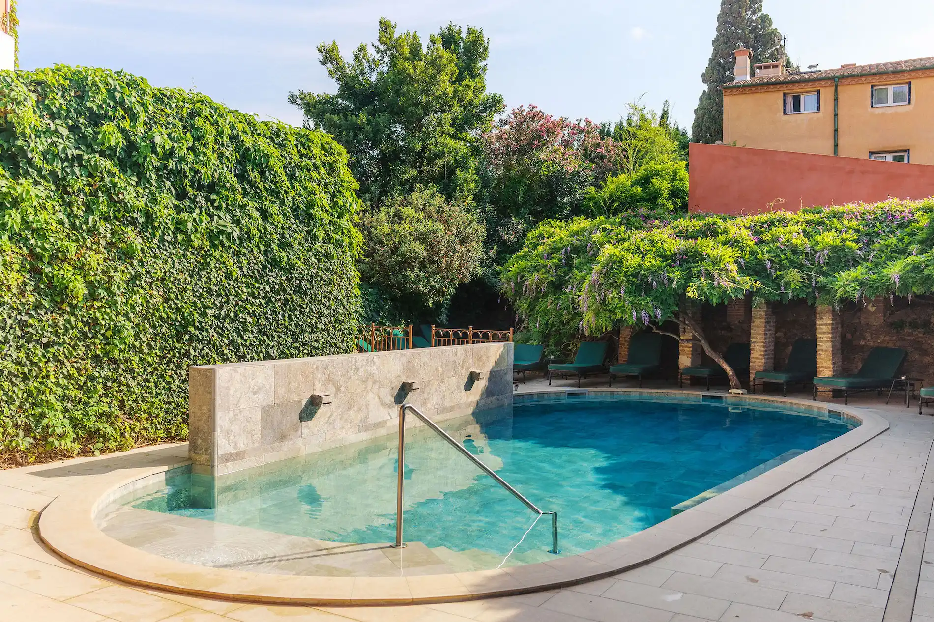 Swimming pool with clear blue water bordered by a tiled patio, a stone water feature wall, and a green hedge on the left; several green lounge chairs sit under a flowering vine-covered pergola with a yellow building in the background.