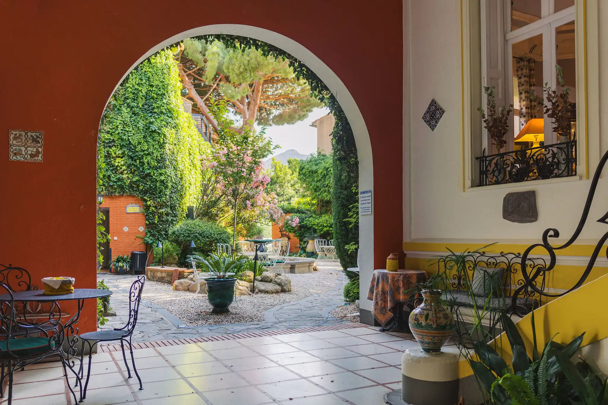 Cozy courtyard with a large arched opening framed by red and white walls, wrought iron chairs and tables, lush green plants, and a stone fountain in the center, surrounded by trees and flowering bushes.